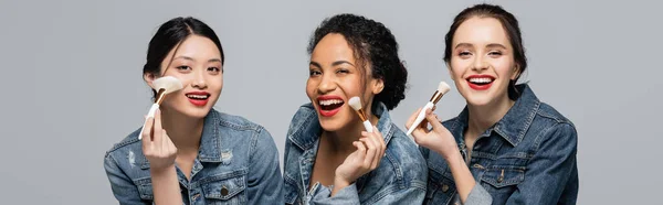 Pretty multiethnic women with red lips holding cosmetic brushes and looking at camera isolated on grey, banner — Stock Photo