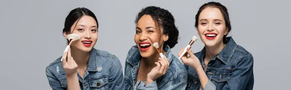 Mujeres bastante multiétnicas con labios rojos sosteniendo cepillos cosméticos y mirando a la cámara aislada en gris, pancarta - foto de stock