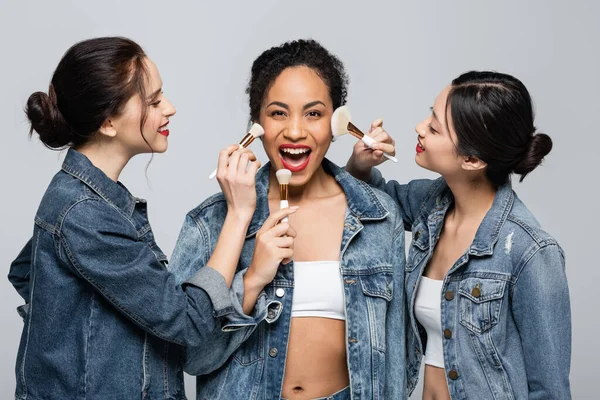 Sonrientes mujeres interracial sosteniendo cepillos cosméticos cerca alegre amigo afroamericano aislado en gris - foto de stock