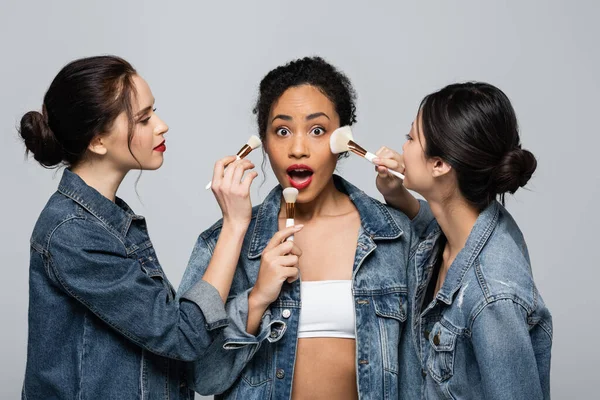 Jeunes femmes interraciales en vestes en denim tenant des pinceaux cosmétiques près d'un ami afro-américain choqué isolé sur gris — Photo de stock