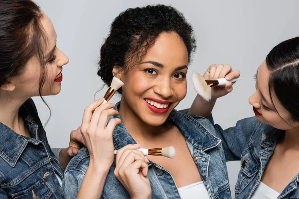 Femme afro-américaine souriante veste en denim debout près d'amis interracial avec des pinceaux cosmétiques isolés sur gris — Photo de stock