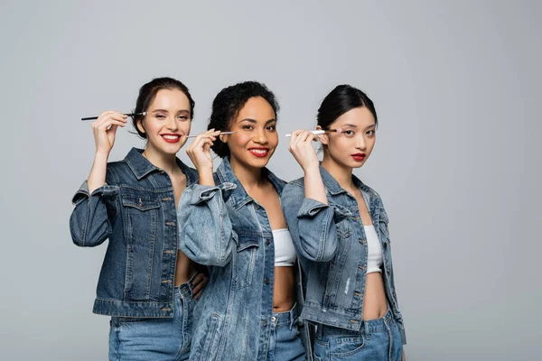 Mulheres inter-raciais alegres em jaquetas jeans segurando escovas cosméticas perto de olhos isolados em cinza — Fotografia de Stock