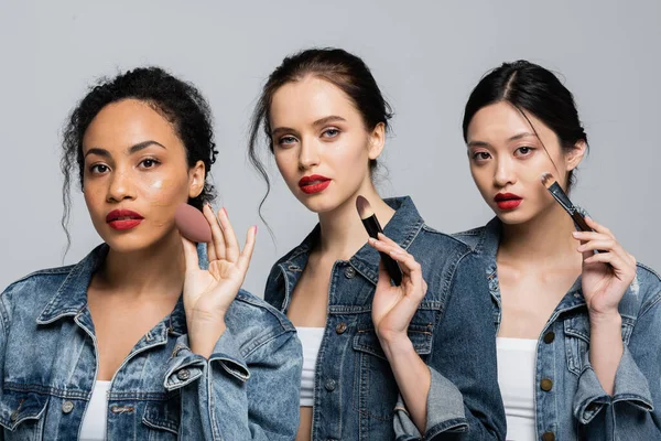 Young multiethnic women in denim jackets holding cosmetic brushes and beauty blender isolated on grey — Stock Photo