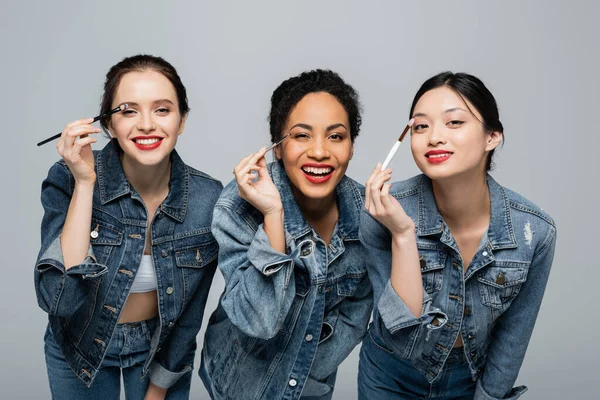 Happy interracial women with red lips holding cosmetic brushes isolated on grey — Stock Photo