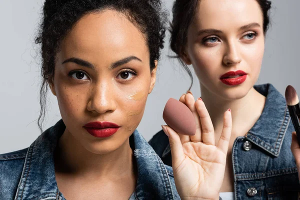 Afroamerikanerin mit Gesichtsstiftung hält Schönheitsmixer in der Nähe Freund mit kosmetischen Pinsel isoliert auf grau — Stockfoto