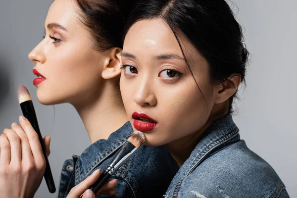 Femme asiatique avec lèvres rouges et fond de teint tenant pinceau cosmétique ami proche sur fond gris — Photo de stock