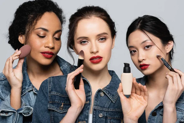 Young woman holding face foundation and cosmetic brush near interracial friends isolated on grey — Stock Photo