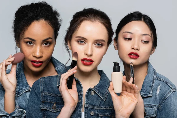 Mujeres interraciales con labios rojos que sostienen cepillos cosméticos y cimientos faciales aislados en gris - foto de stock