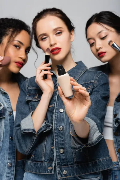 Fundación de la cara en la mano de la mujer borrosa cerca de amigos interracial con cepillos cosméticos aislados en gris - foto de stock