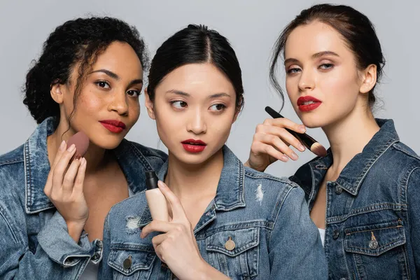 Interracial young women holding cosmetic brush, face foundation and beauty blender isolated on grey — Stock Photo