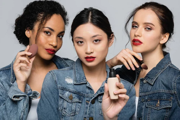 Asian woman holding face foundation near interracial friends with beauty blender and cosmetic brush isolated on grey — Stock Photo