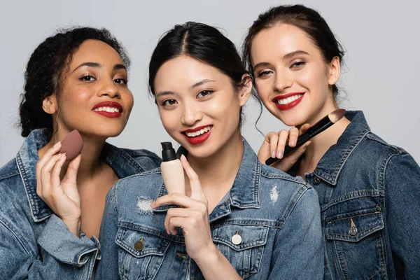 Sonriente asiático mujer holding cara fundación cerca interracial amigos con rojo labios aislado en gris - foto de stock