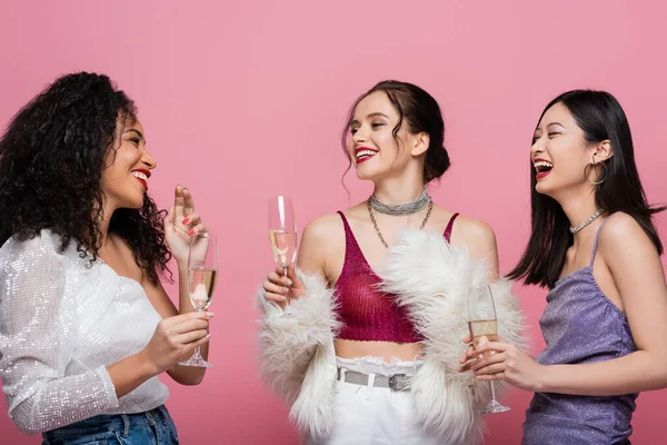 Улыбчивые и стильные межрасовые женщины, держащие в руках шампанское и разговаривающие изолированно на розовом — стоковое фото