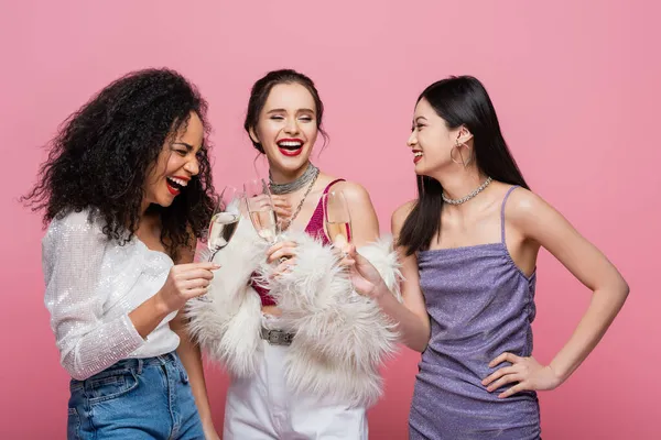Смеющиеся мультикультурные друзья, держащие шампанское во время вечеринки, изолированные от розового — стоковое фото