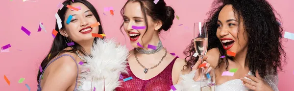 Cheerful multicultural women holding champagne under confetti on pink background, banner — Stock Photo