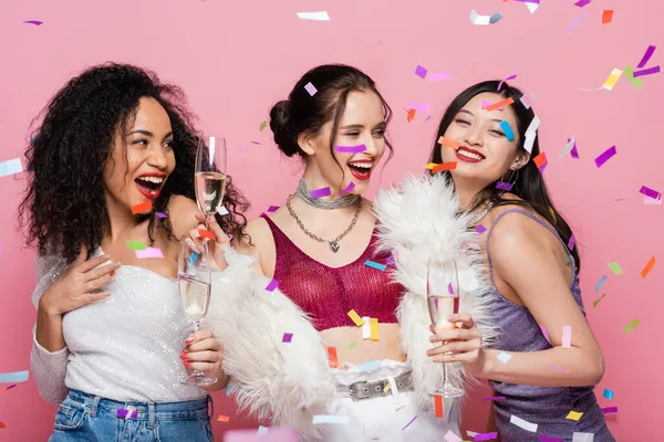 Mulheres multiétnicas elegantes segurando copos de champanhe sob confete no fundo rosa — Fotografia de Stock