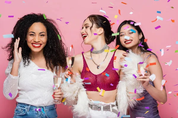 Femmes multiethniques branchées avec champagne debout sous confettis sur fond rose — Photo de stock