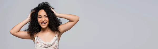 Sonriente mujer afroamericana tocando el cabello aislado en gris, pancarta - foto de stock