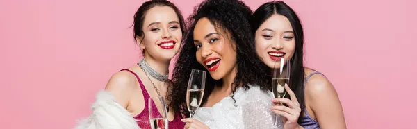 Sonrientes mujeres interracial celebración de champán cerca de elegante amigo afroamericano aislado en rosa, bandera - foto de stock