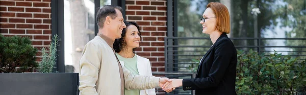 Happy realtor in glasses shaking hands with man standing near african american wife and modern house, banner — Stock Photo