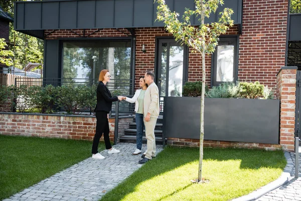 Felice agente immobiliare in occhiali stringendo la mano con donna afro-americana vicino marito e casa moderna — Foto stock