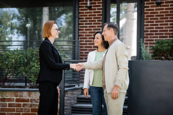 Heureux agent immobilier dans lunettes serrant la main avec l'homme debout près de femme afro-américaine et maison moderne — Photo de stock