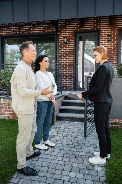 Alegre agente inmobiliario en gafas mirando interracial pareja cerca de casa moderna - foto de stock