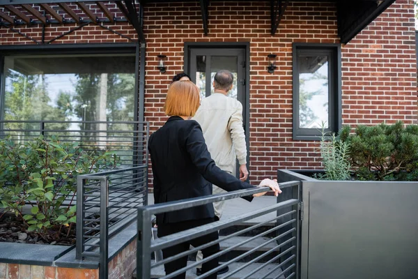 Back view of redhead realtor walking behind couple while entering modern house — Stock Photo