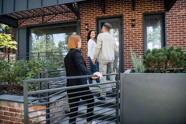 Rousse agent immobilier debout derrière couple interracial près de maison moderne — Photo de stock