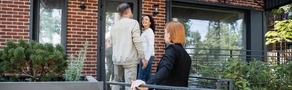 Redhead realtor in piedi dietro la coppia interrazziale vicino alla casa moderna, banner — Foto stock