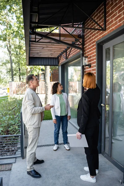 Full length of redhead realtor showing new modern house to multiethnic couple — Stock Photo