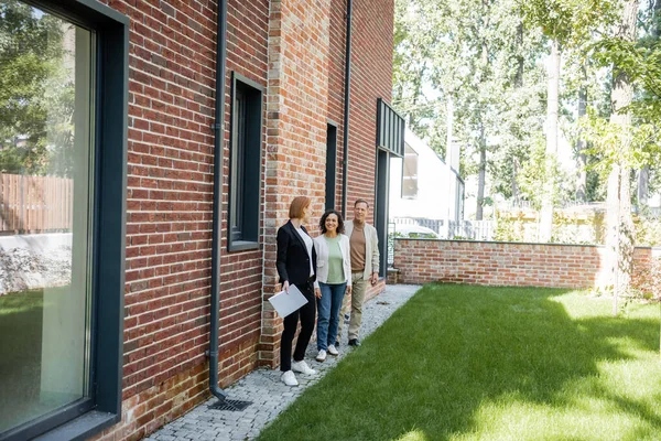 Longitud completa de pelirroja agente inmobiliario en gafas celebración carpeta mientras mira feliz pareja multiétnica cerca de casa - foto de stock