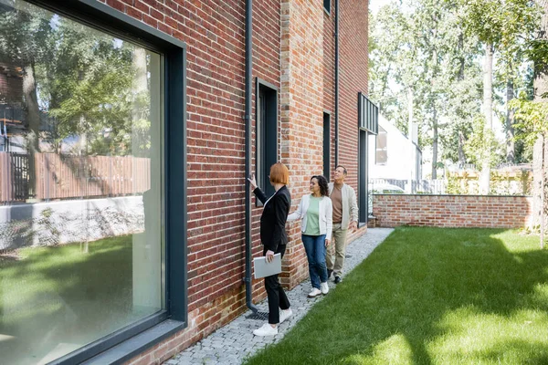 Full length of redhead realtor holding folder while showing house to happy multiethnic couple — Stock Photo