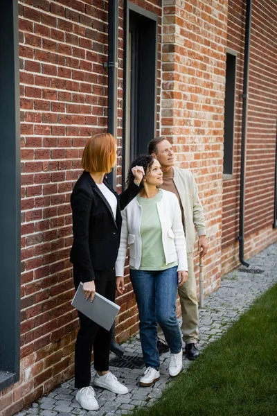 Pleine longueur de rousse agent immobilier tenant dossier tout en montrant la maison au couple multiethnique — Photo de stock