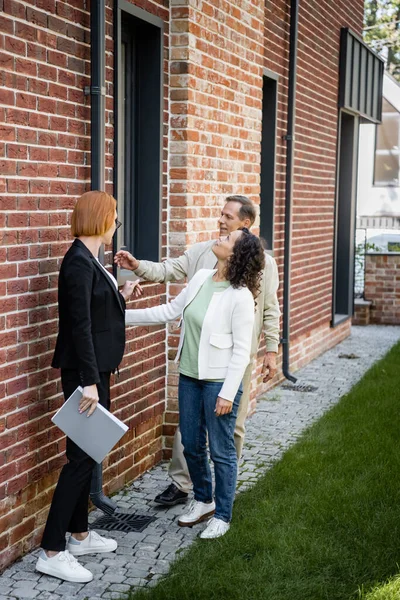 Toute la longueur du dossier de tenue de l'agent immobilier tout en montrant la maison au couple multiethnique — Photo de stock