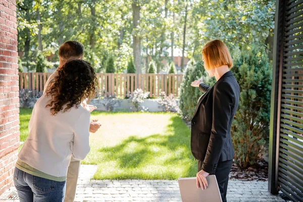 Back view of redhead realtor in glasses pointing at lawn to couple — Stock Photo