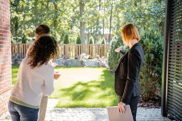 Vue arrière de rousse agent immobilier dans des lunettes pointant vers la pelouse à couple — Photo de stock