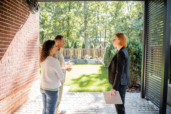 Redhead realtor in glasses talking with interracial couple — Stock Photo