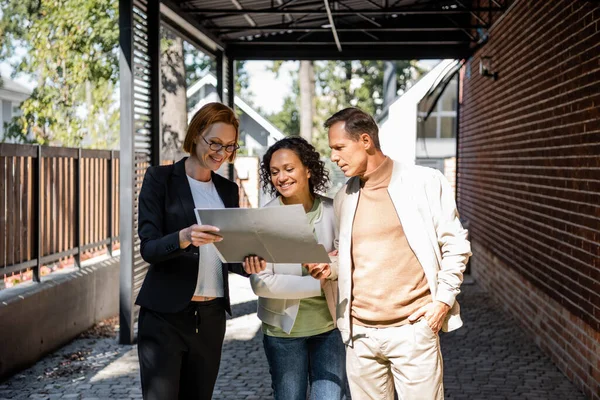 Happy redhead realtor in glasses showing folder to cheerful interracial couple — Stock Photo