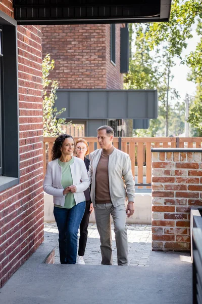 Alegre afroamericana mujer caminando cerca de marido y agente inmobiliario fuera - foto de stock