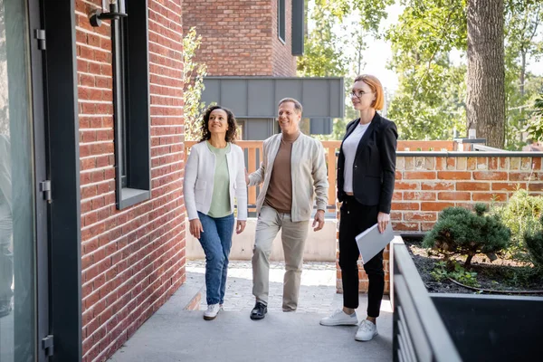 Redhead realtor in glasses holding folder while looking at new house near cheerful interracial couple — Stock Photo