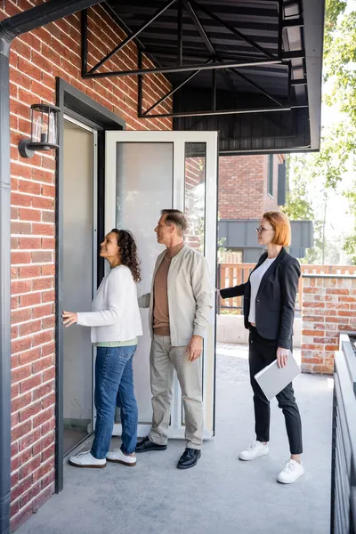 Agente immobiliare in occhiali apertura porta mentre coppia interrazziale entrare nuova casa — Foto stock