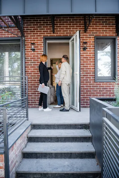 Redhead realtor in glasses pointing with finger at house while talking with interracial couple — Stock Photo
