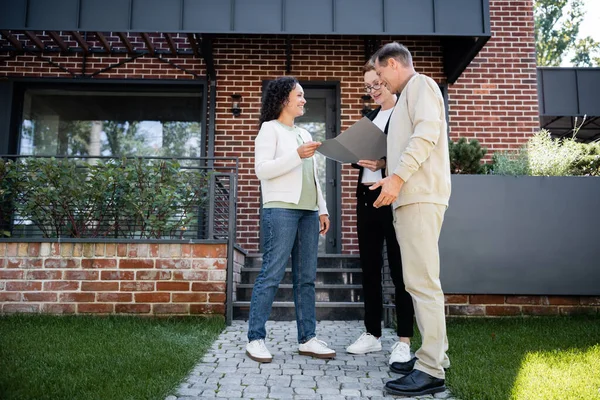 Happy multiethnic couple looking at folder near realtor and modern house outside — Stock Photo