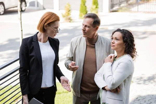 Redhead realtor with folder gesturing near husband and african american wife — Stock Photo