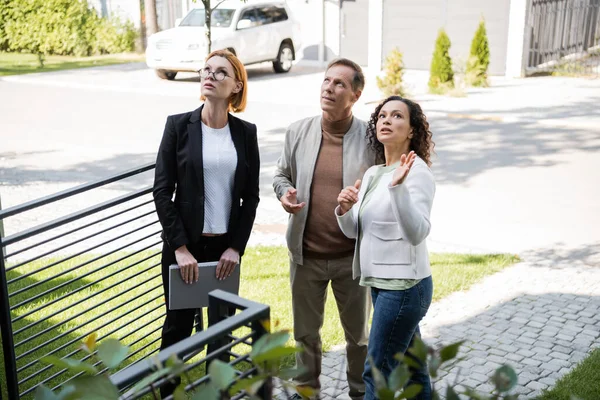 Redhead realtor with folder looking away near man and african american woman pointing away — Stock Photo