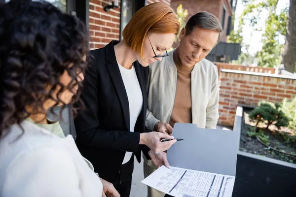 Rousse agent immobilier tenant stylo et dossier avec demande de prêt près de couple multiethnique — Photo de stock