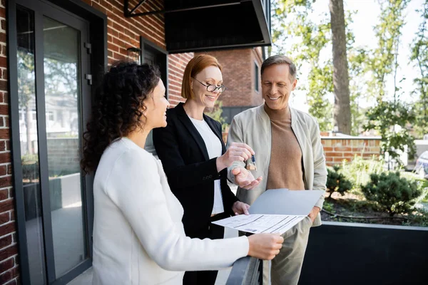 Happy realtor giving key to cheerful man near african american wife — Stock Photo