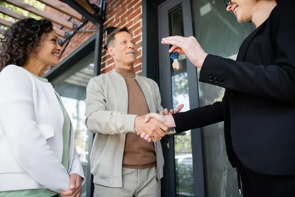 Low angle view of happy realtor holding key while shaking hands with cheerful man near african american wife — Stock Photo