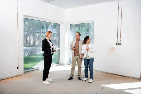 Pelirroja agente inmobiliario en gafas celebración de tableta digital cerca de pareja interracial dentro de la nueva casa - foto de stock